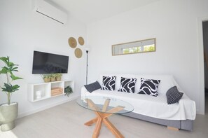 Ein modernes Wohnzimmer mit einem weißen Sofa, einem Glastisch, einem an der Wand montierten Fernseher und einer kleinen Pflanze.