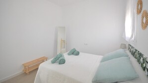 A minimalist bedroom with a bed, a bench, a mirror, and a window with a curtain. - Holiday Home 'Casa Turquesa' with Private Pool, Wi-Fi and Air Conditioning (Conil de la Frontera)