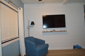 A room with a blue sofa, a mounted flat-screen TV, a lamp, and a shelf with decorative items.
