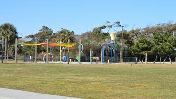 An outdoor park with playground equipment, a covered seating area, and trees in the background.
