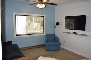 A living room with a ceiling fan, a large window with blinds, a blue armchair, a flat-screen TV on a wall-mounted shelf, and a dark-colored sofa. - Small Peace of Paradise,  In beautiful Oak Island (Oak Island)