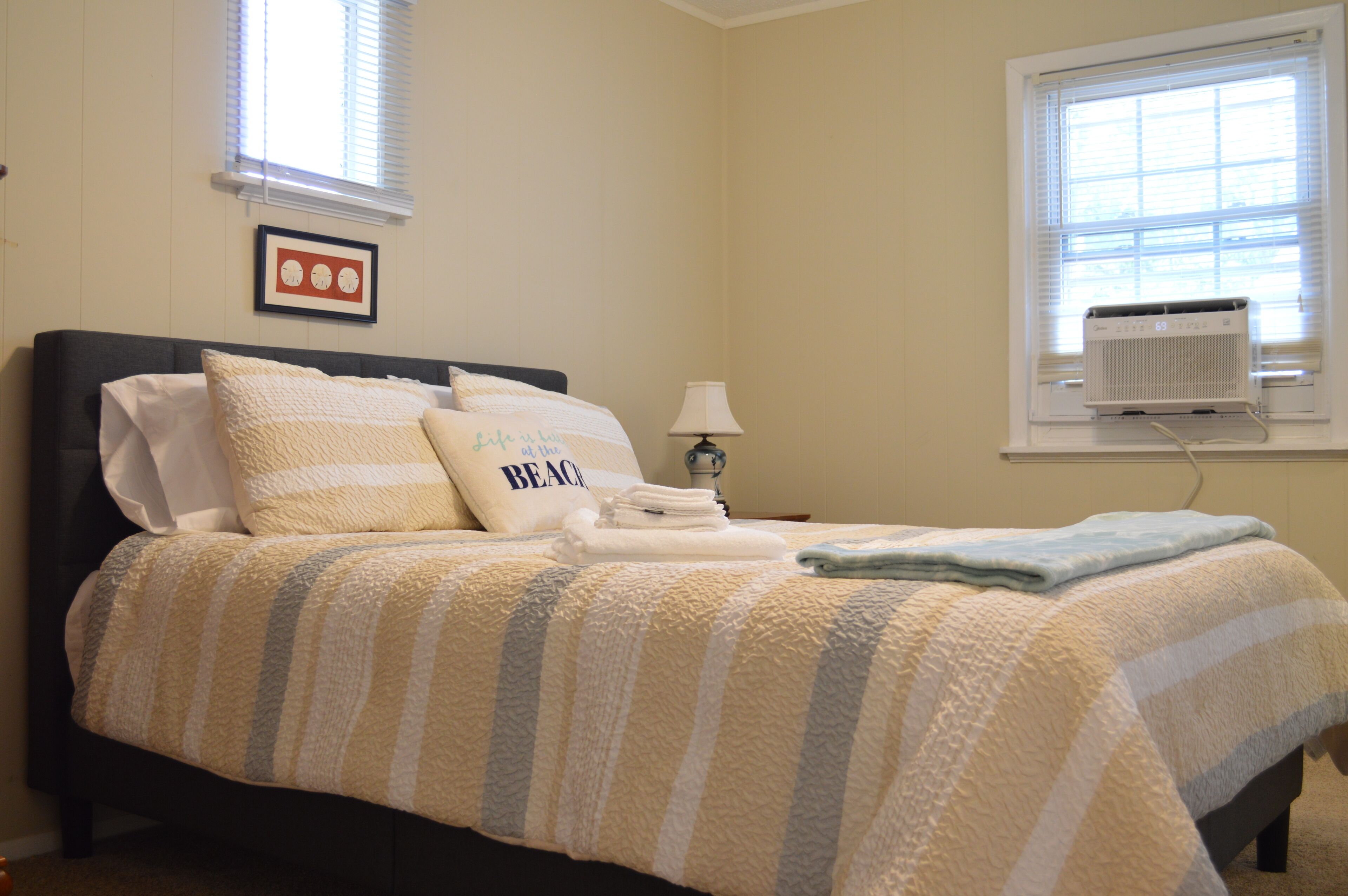 Una cama bien tendida con ropa de cama a rayas, un almohadón con la palabra 'BEACH' y un aire acondicionado de pared.