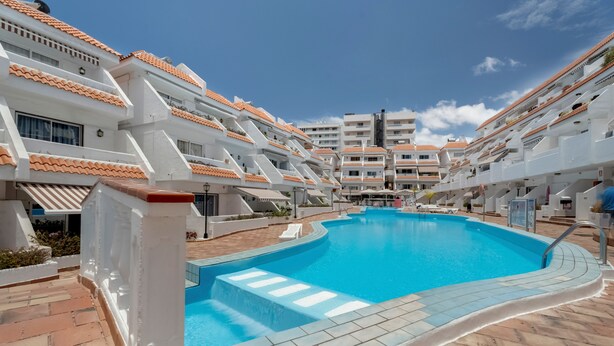 Une piscine entourée de bâtiments blancs aux toits recouverts de tuiles orange, sous un ciel bleu clair, et bordée d’une terrasse carrelée.