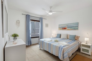 A bedroom with a bed, bedside tables, a ceiling fan, a window with curtains, a wall painting, and a small plant on a dresser.