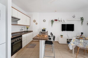 A modern kitchen with white cabinets, a granite countertop, a built-in oven, a dining area with a table and chairs, and a flat-screen TV mounted on the wall.
