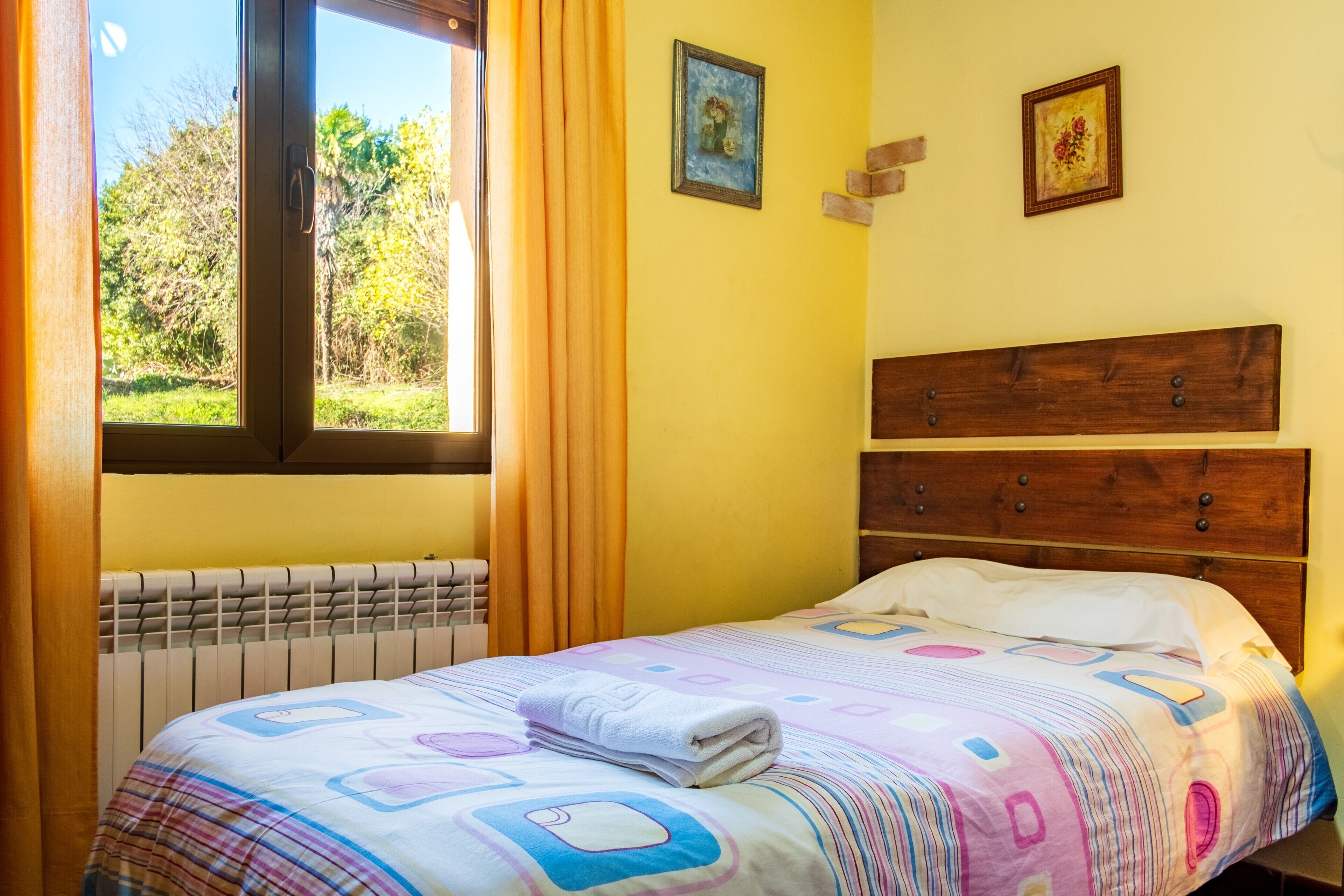 Un dormitorio con una cama, una ventana con cortinas, un radiador y dos cuadros en la pared.