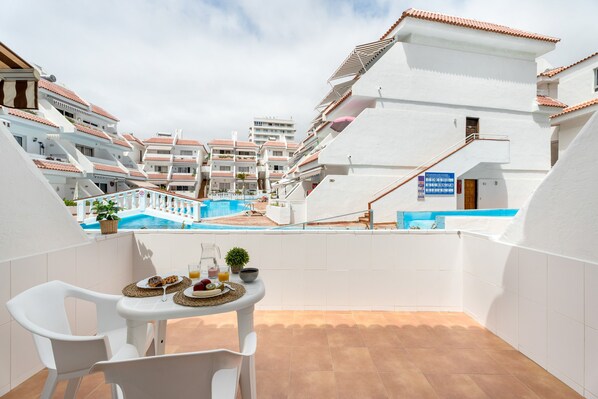 A balcony with white chairs and a table set with breakfast, overlooking a swimming pool and adjacent buildings.