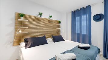 Una cama doble con dos almohadas, una mesita de noche con una lámpara, un estante en la pared con plantas y una cortina azul.