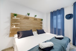 A double bed with two pillows, a bedside table with a lamp, a wall-mounted shelf with plants, and a blue curtain.