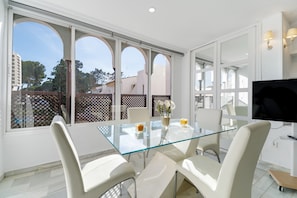A modern dining area with a glass table, white chairs, and a TV.