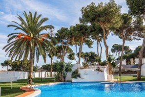 A swimming pool surrounded by palm trees and pine trees, with a white building in the background.