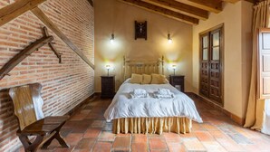 Una habitación con pared de ladrillo, vigas de madera, una cama con ropa blanca, dos mesitas de noche con lámparas y un banco de madera.