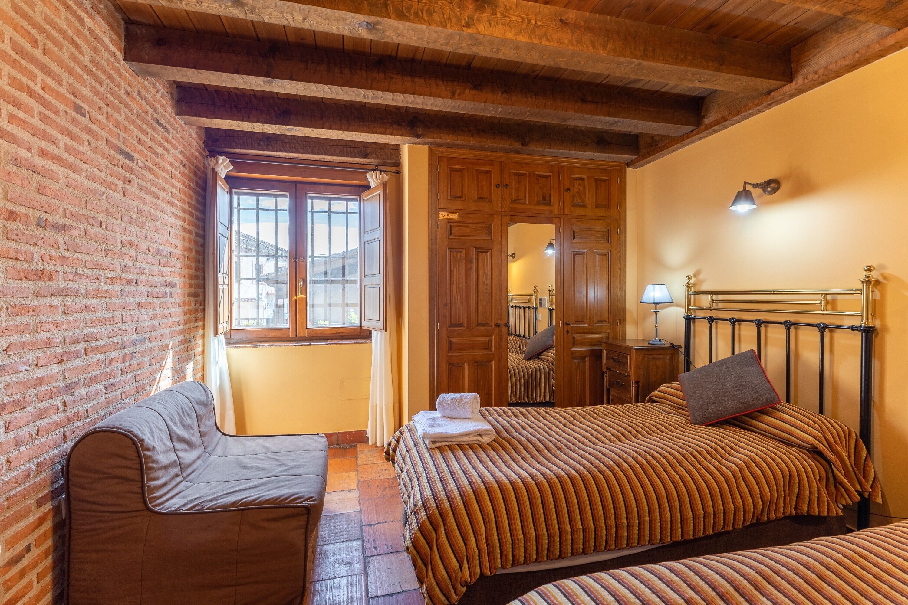 Een kamer met een bakstenen muur, houten balken in het plafond, een bed met gestreept beddengoed, een bank en een raam met gordijnen.