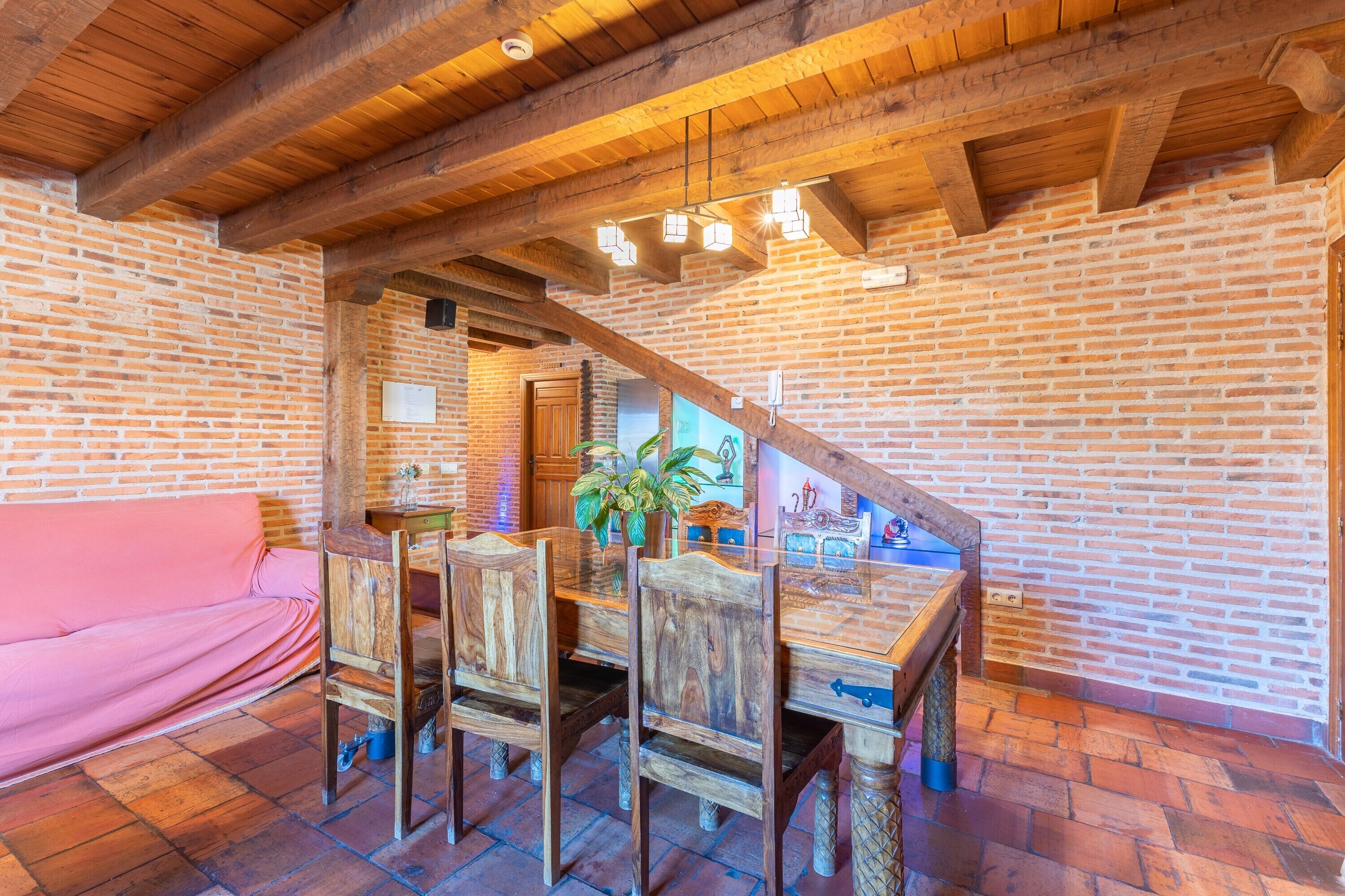 Un interior rústico con vigas de madera a la vista, pared de ladrillo, mesa de comedor con sillas y sofá rosa.