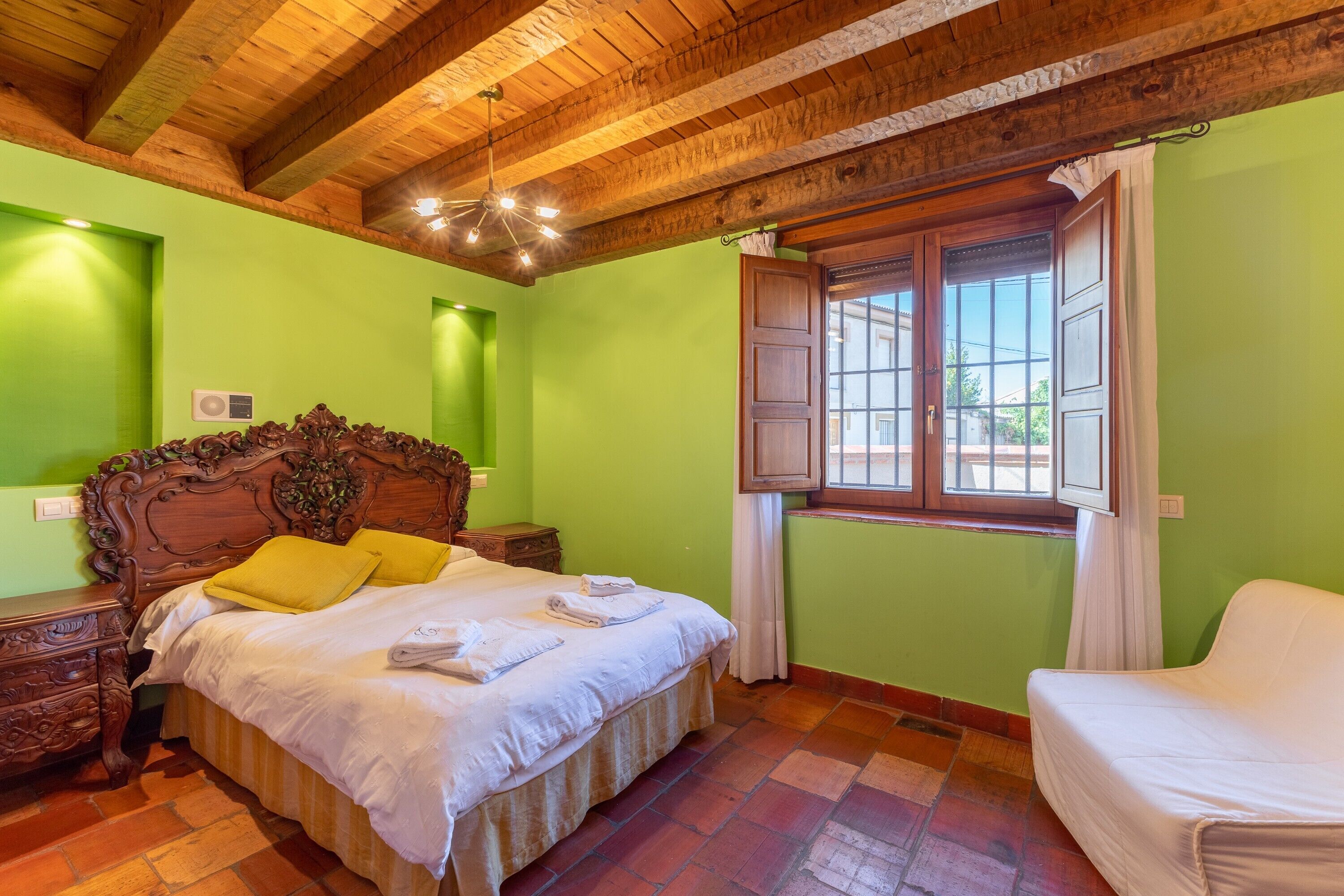 A room with a wooden ceiling, a bed with a headboard, a window with curtains, and a white chair.