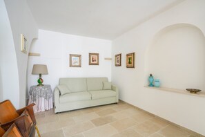 A living room with a sofa, a small table with a lamp, framed pictures on the wall, and a shelf with decorative items.