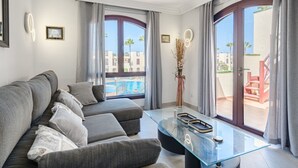 A modern living room with a glass coffee table, grey sofa, and large windows offering a view of a pool and buildings.