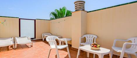 Una terraza en la azotea con sillas de plástico blanco y una mesa, una mesita con una cesta de frutas, y un cielo azul despejado.
