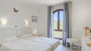 A bedroom with a bed, bedside tables, a chair, a desk, a window with curtains, and a wall-mounted light fixture.