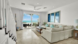 Een ruime woonkamer met een groot raam met uitzicht op het strand, een plafondventilator en een beige bank met teal kussens.