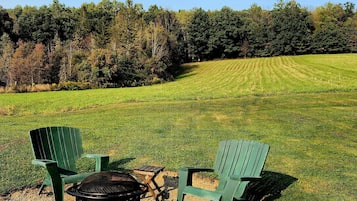 Two green Adirondack chairs and a black grill in a grassy field with a forest in the background.