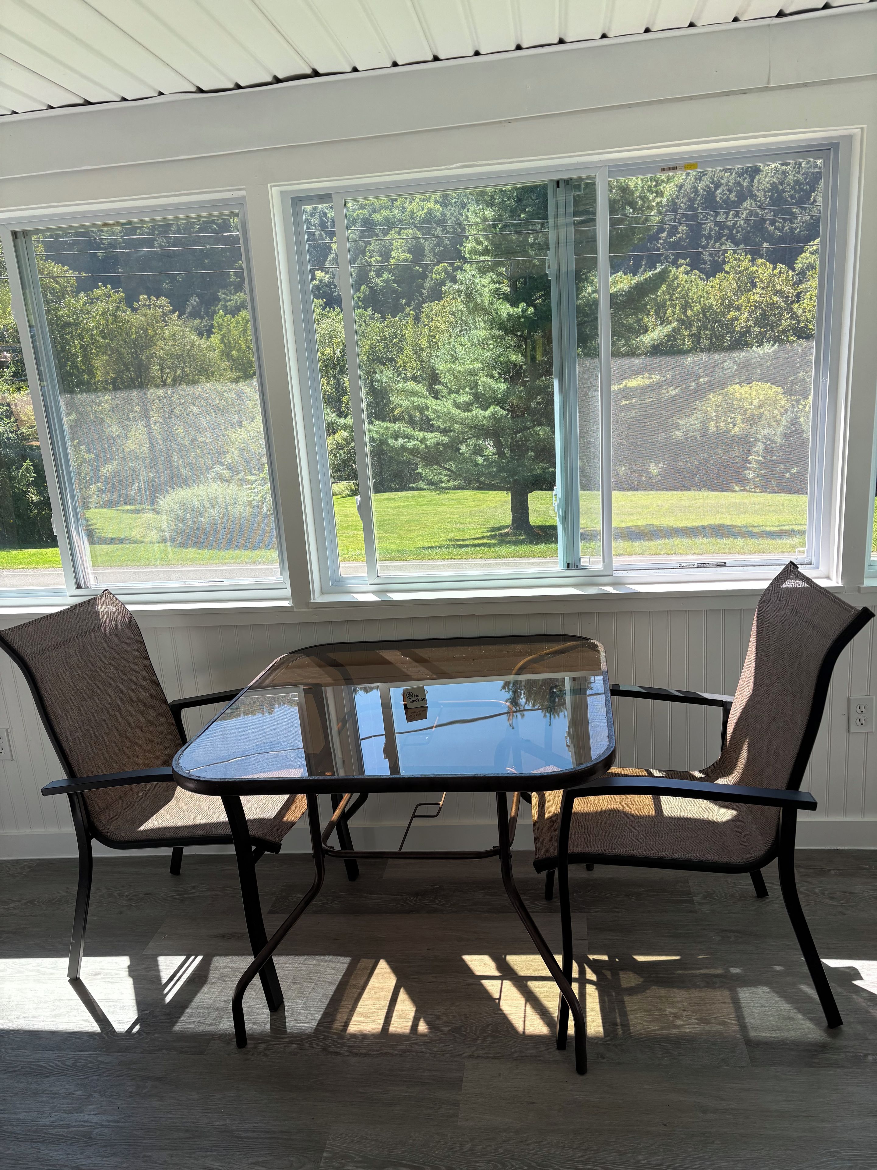 A room with a glass table and two chairs, large windows with a view of greenery, and a white ceiling.