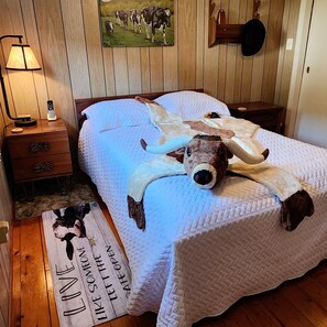 A bedroom with a bed, a lamp, a nightstand, a drawer, a painting, and a rug with text. - 50 Acres of Adventure: Discover Walter’s Place” (Cameron)