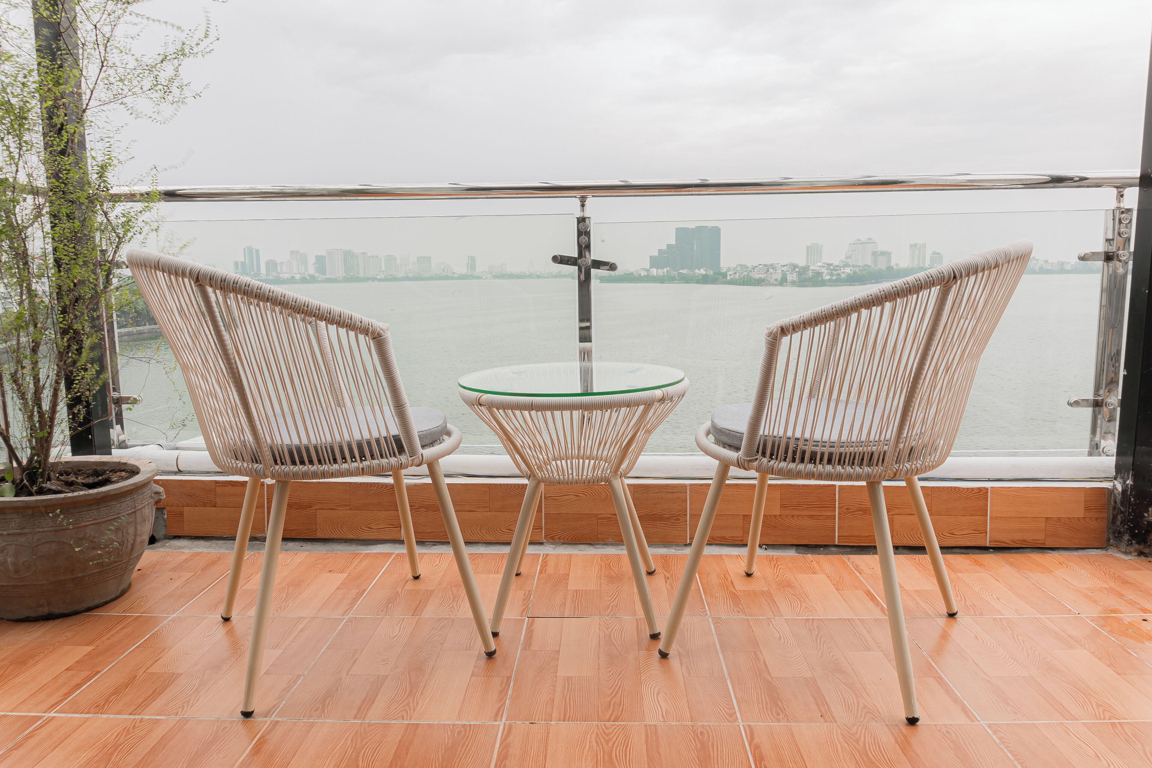 Un balcón con dos sillas de mimbre y una mesa de vidrio, con vista a una masa de agua y al horizonte de una ciudad.