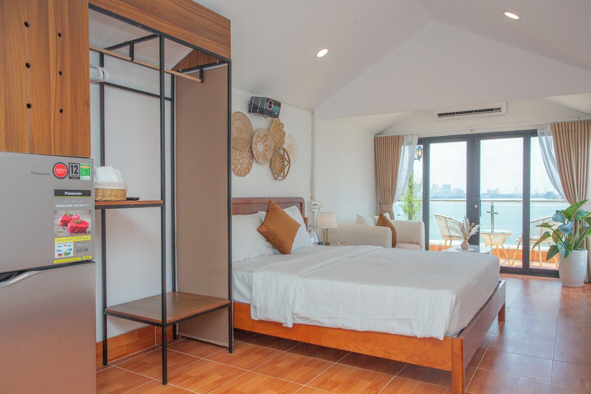 Een hotelkamer met een bed, een houten kledingkast, uitzicht op zee en een balkon met stoelen.