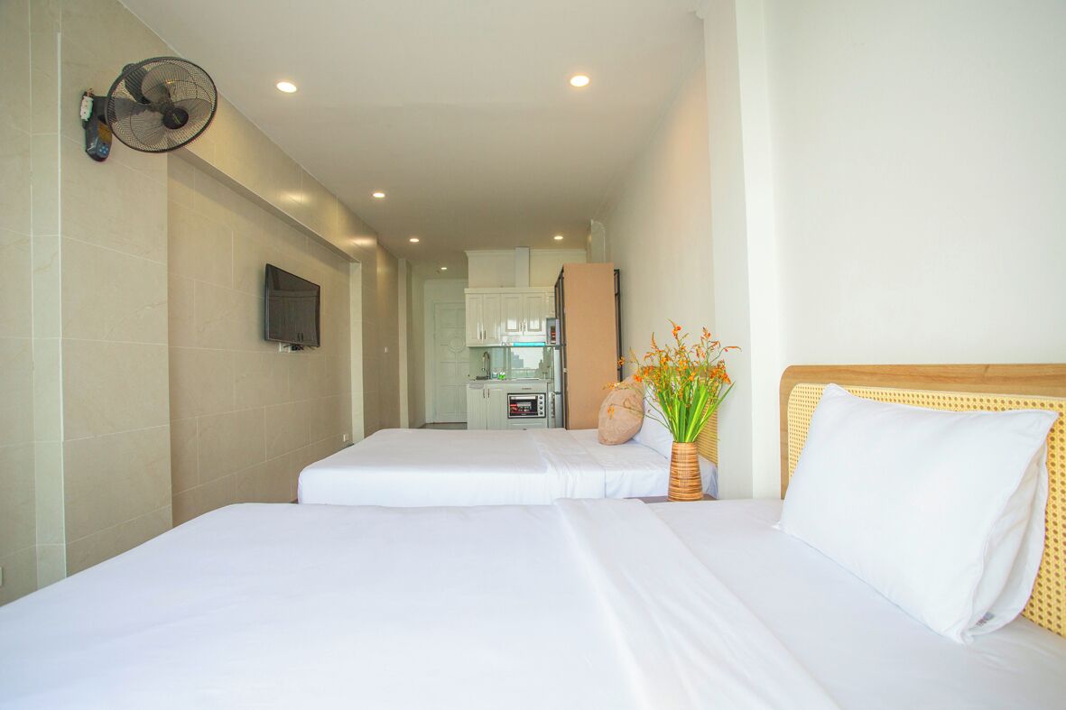 Habitación de hotel con dos camas, televisión montada en la pared, ventilador, pequeña cocina y un florero decorativo con flores.
