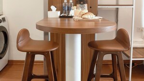 Ein kleiner Holztisch mit zwei braunen Hocker, einem Blumentopf und einigen Gegenständen darauf, darunter eine Flasche, eine Taschentuchbox und ein Croissant.