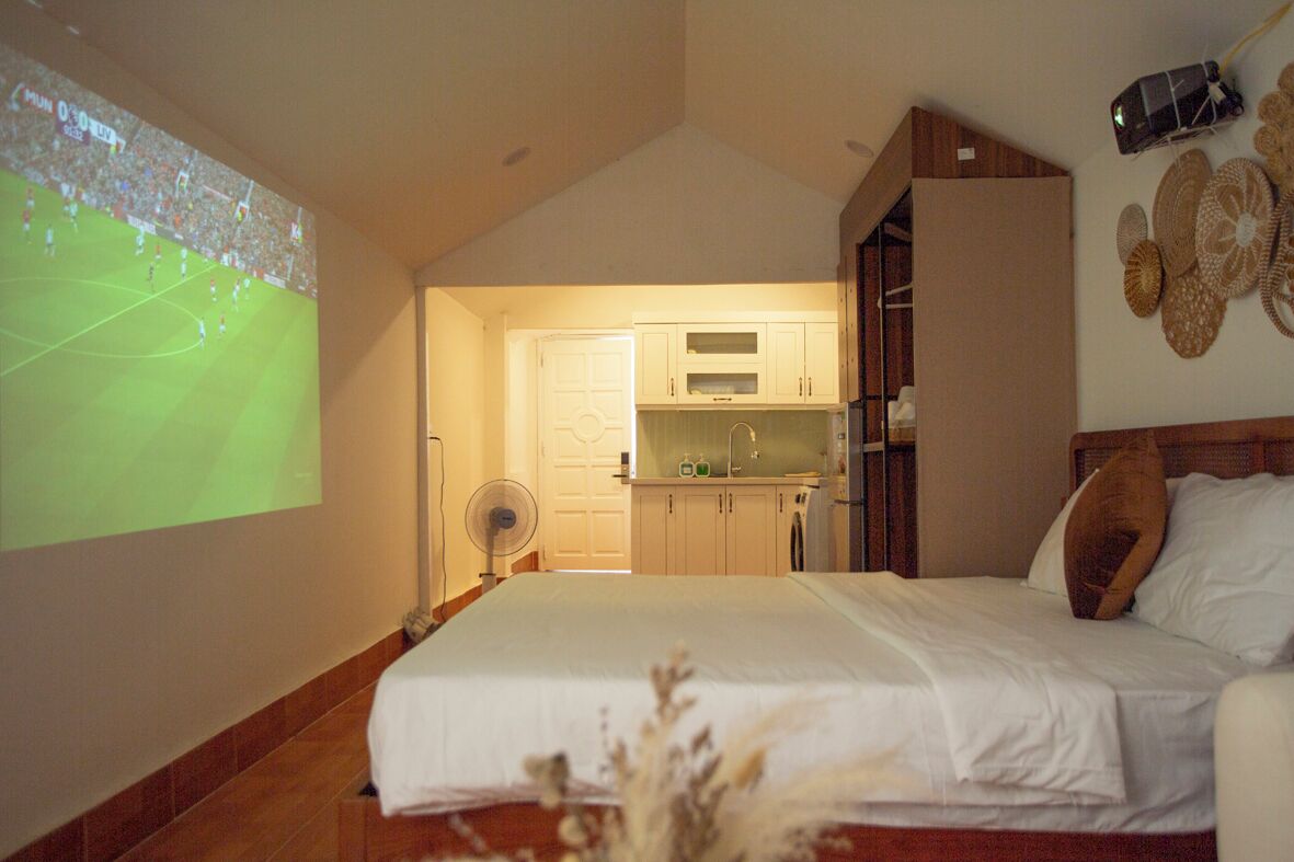 A bedroom with a bed, a fan, and a kitchen in the background.