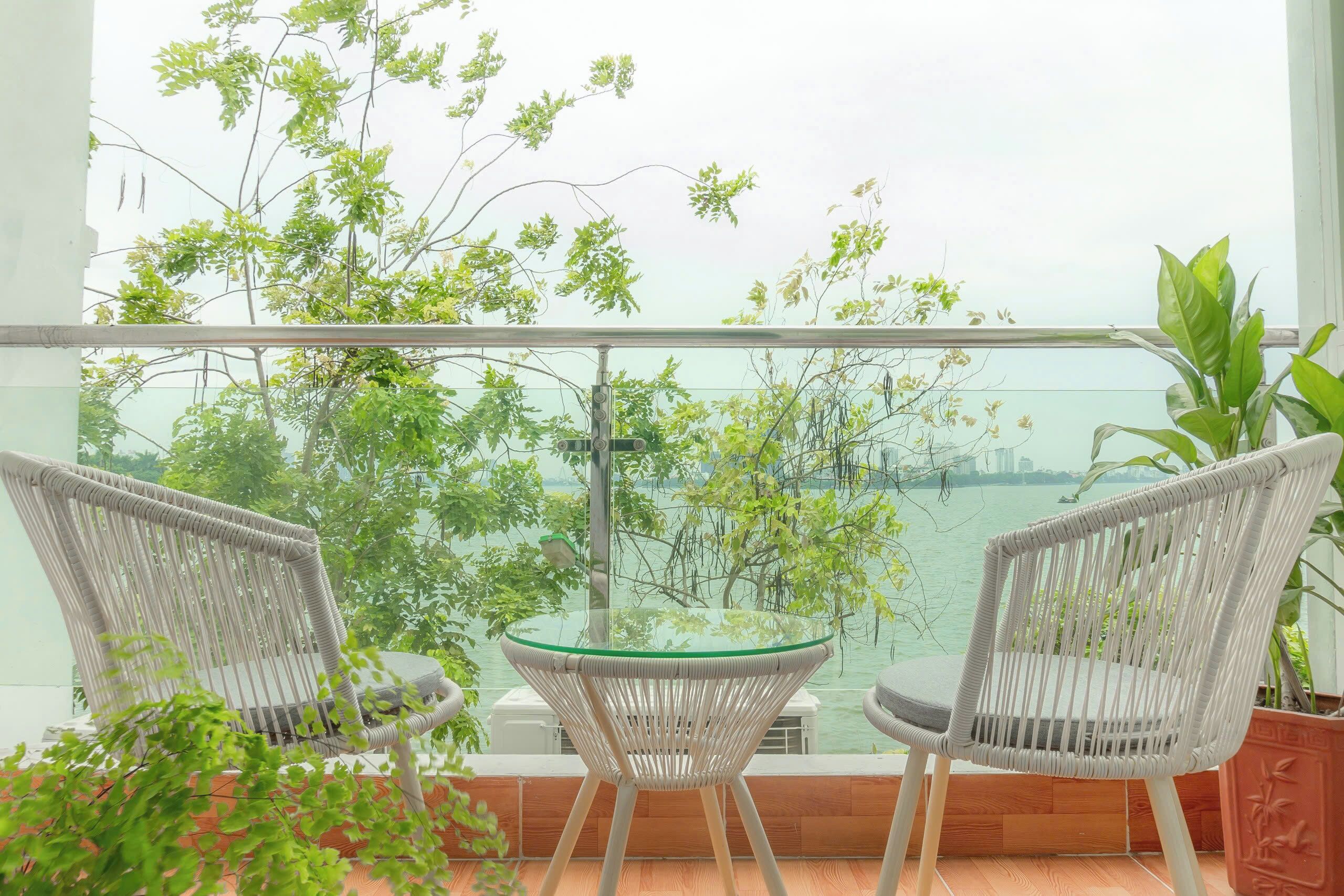 A balcony with two wicker chairs and a glass table, overlooking a body of water and greenery.