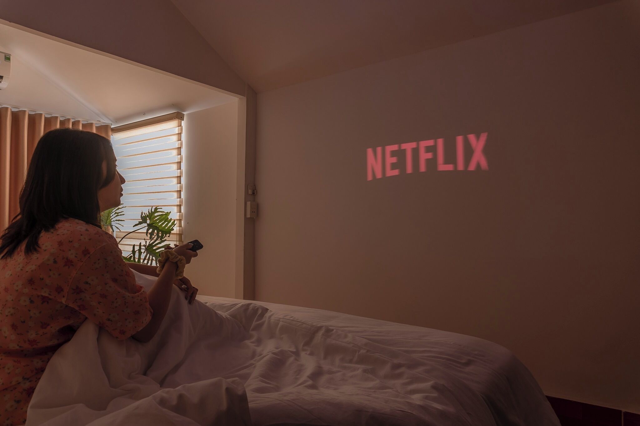 A person sitting on a bed in a room with a wall displaying the Netflix logo.