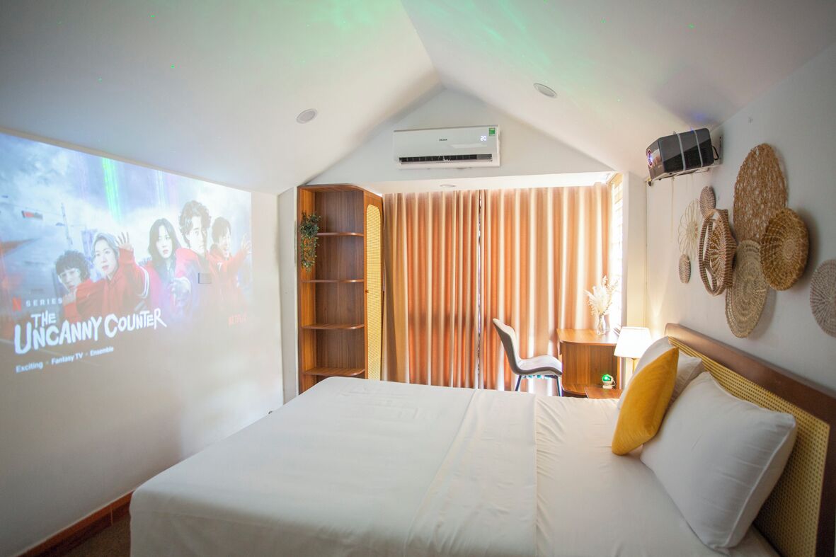 A hotel room with a large bed, a TV screen displaying a show, a bookshelf, a window with curtains, and a wall-mounted speaker.