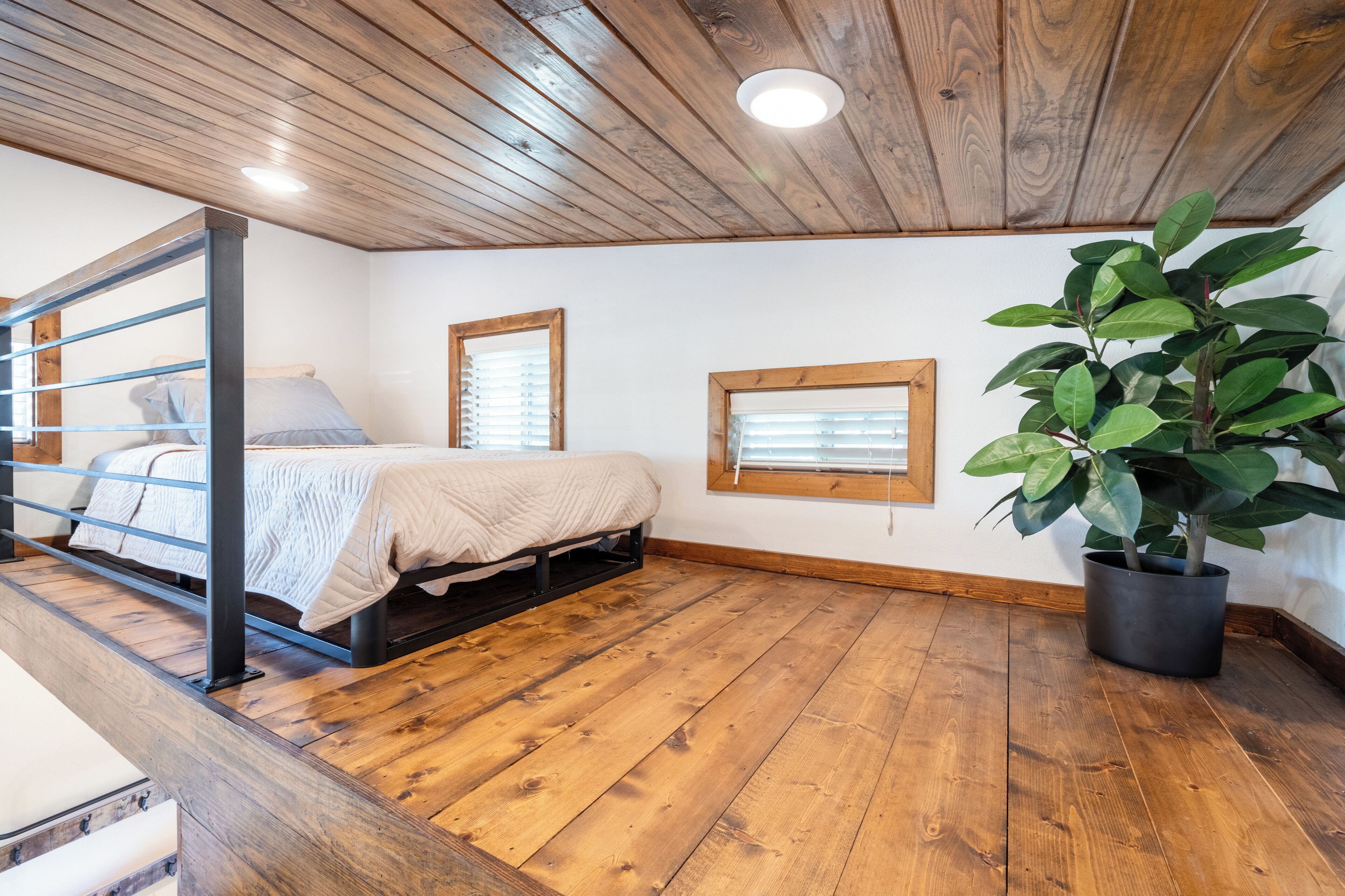 A bedroom with a wooden floor, a bed with a white bedspread, a window with blinds, and a potted plant.