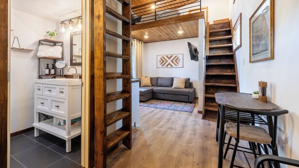 A modern interior with a wooden staircase, a white vanity, a gray sofa, and a dining area with stools. - Charming Tiny Home Cottage - East Side! (Austin)