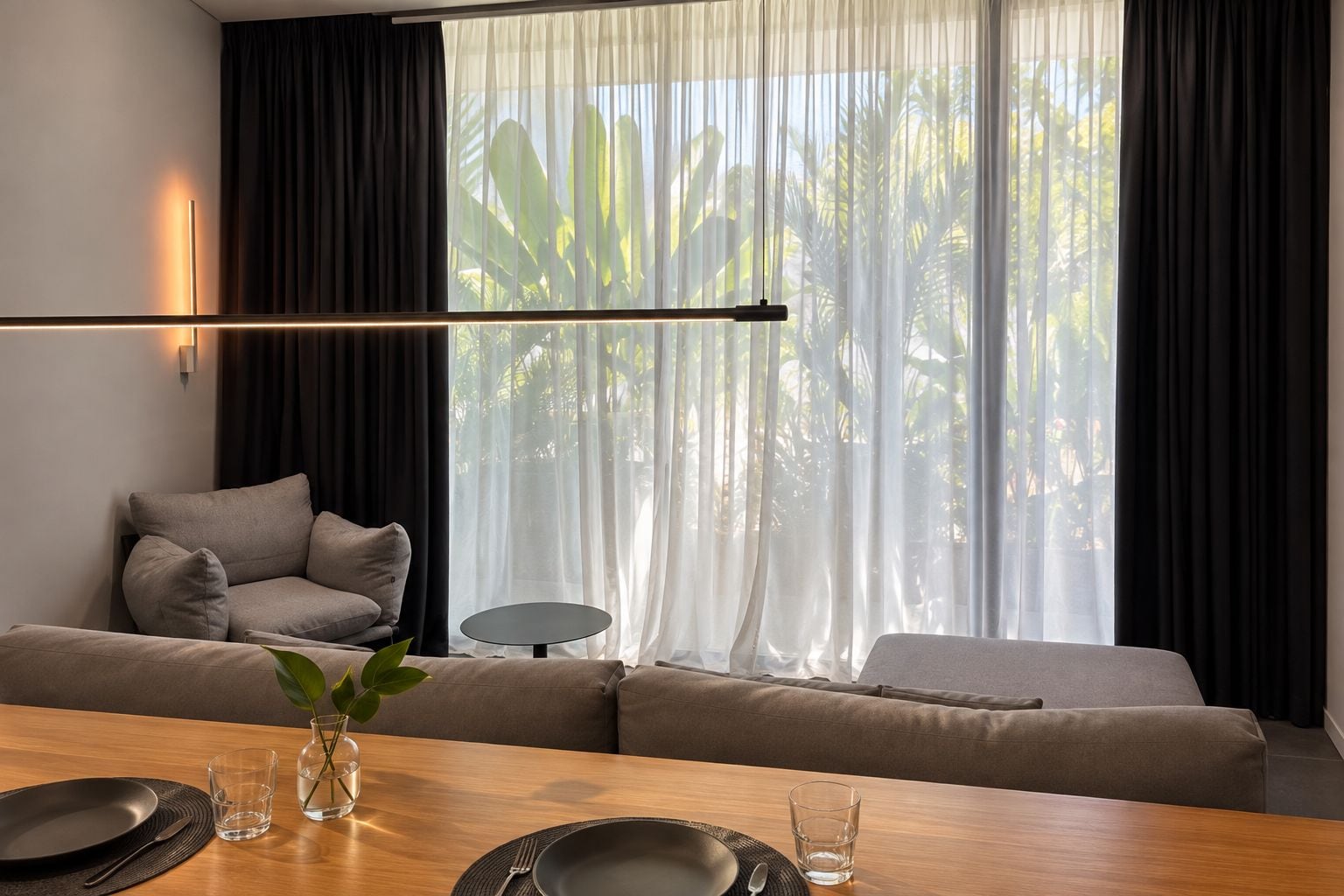 A modern living room with a sofa, a dining table set for two, and a large window with sheer curtains.