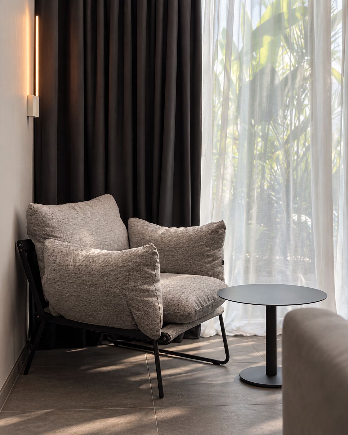 Un fauteuil moderne avec un coussin, placé à côté d’une table ronde dans une pièce dotée d’une grande fenêtre, de rideaux et d’une lampe fixée au mur.
