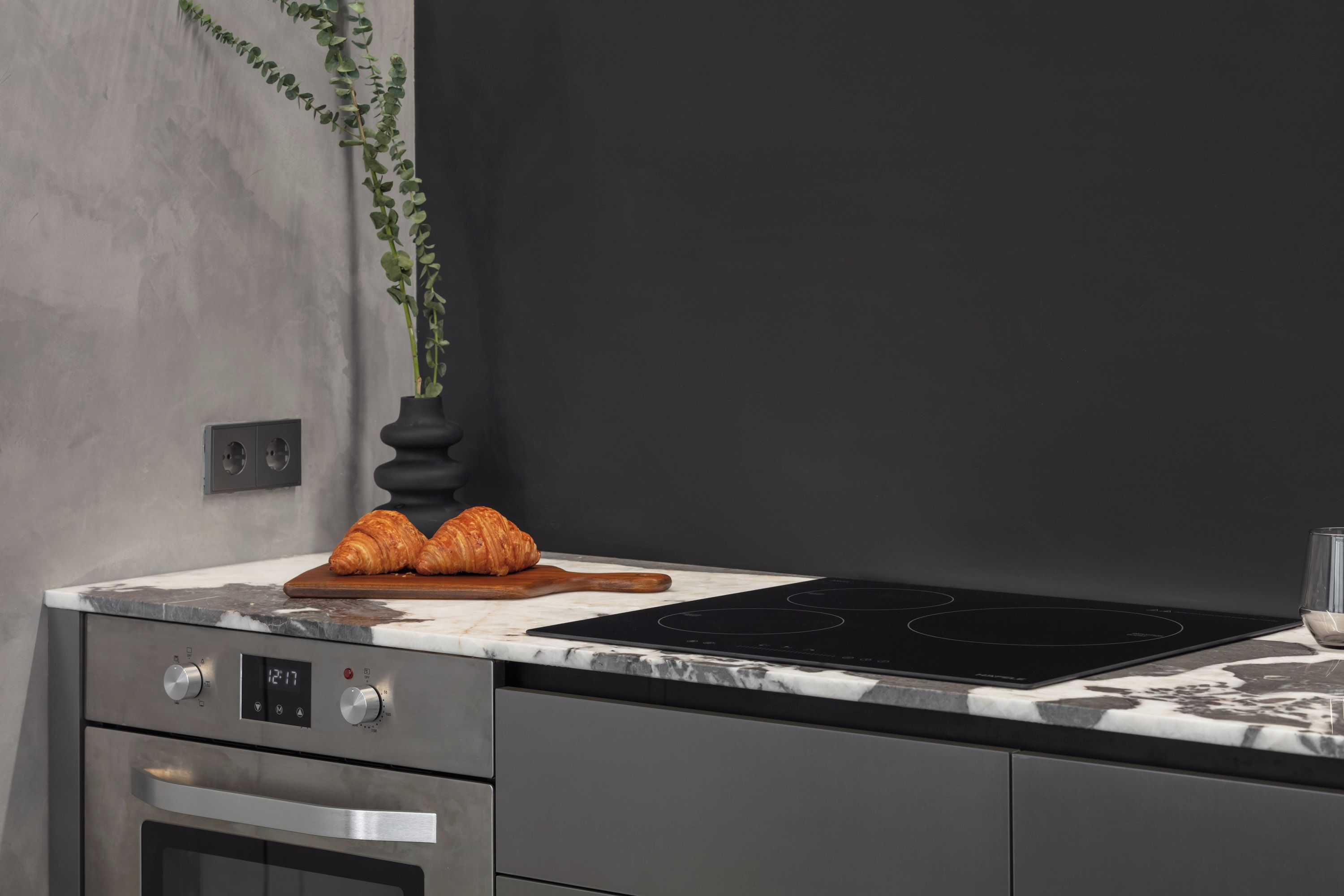 A kitchen with a marble countertop, a black induction cooktop, a stainless steel oven, and a wooden cutting board with croissants.