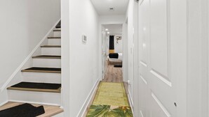 Un couloir étroit, avec un escalier en bois sur la gauche, un tapis à motifs verts sur le sol et une porte blanche sur la droite.