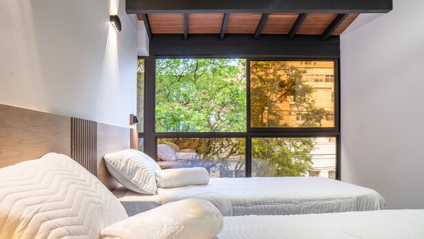 A bedroom with two beds, a large window, and a view of trees and buildings outside.