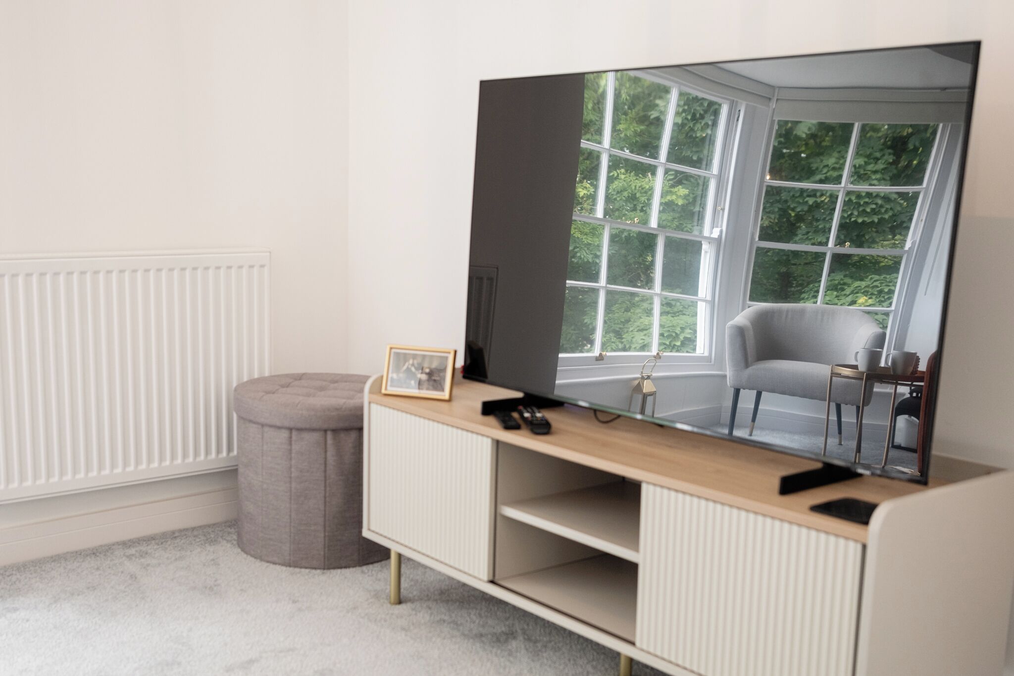 A modern living room with a large flat-screen TV mounted on a wall, a grey ottoman, and a white radiator.