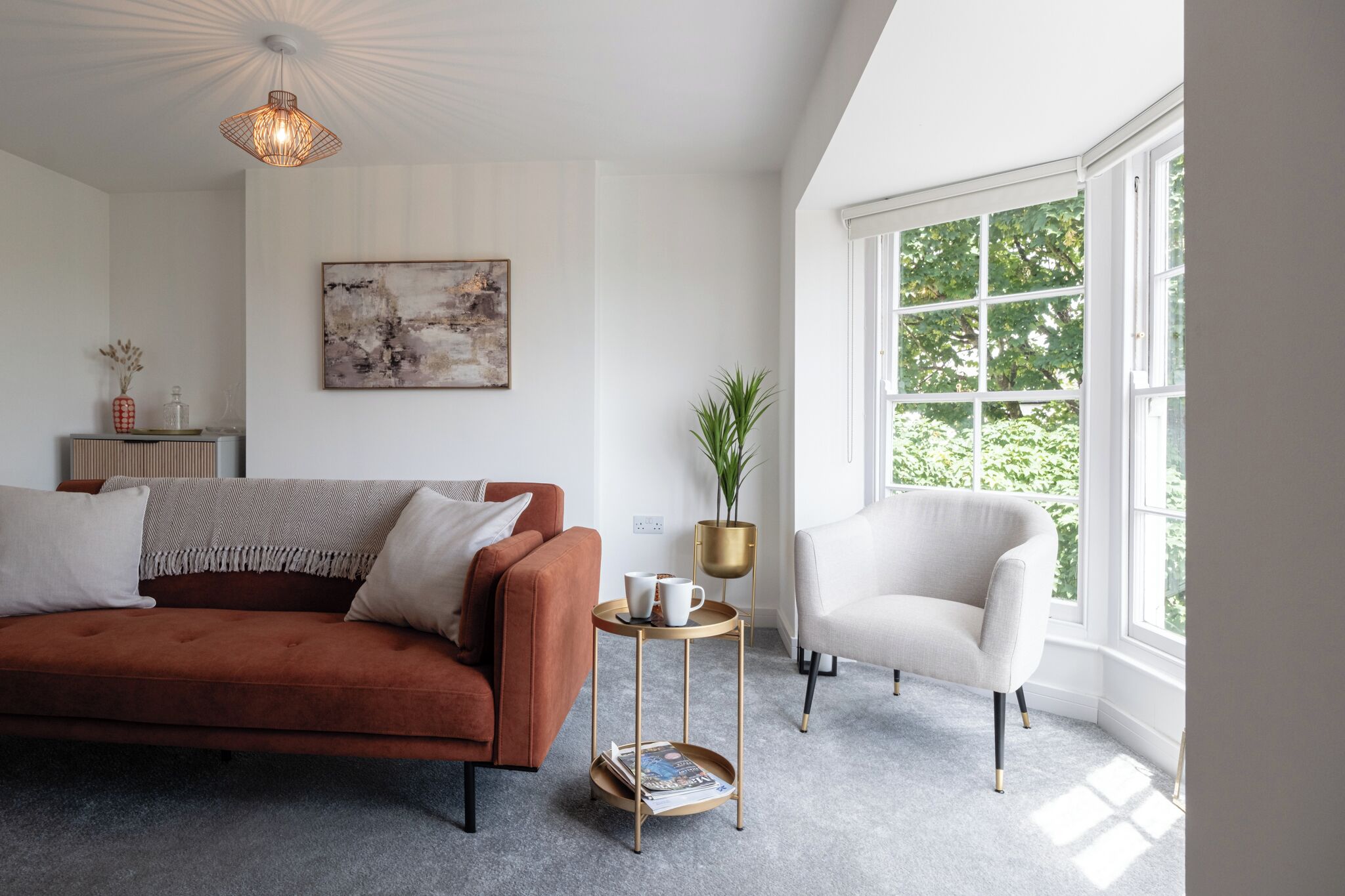 Ein modernes Wohnzimmer mit einem braunen Sofa, einem weißen Sessel, einem runden Beistelltisch mit Zeitschriften und einem großen Fenster mit Blick auf Bäume.