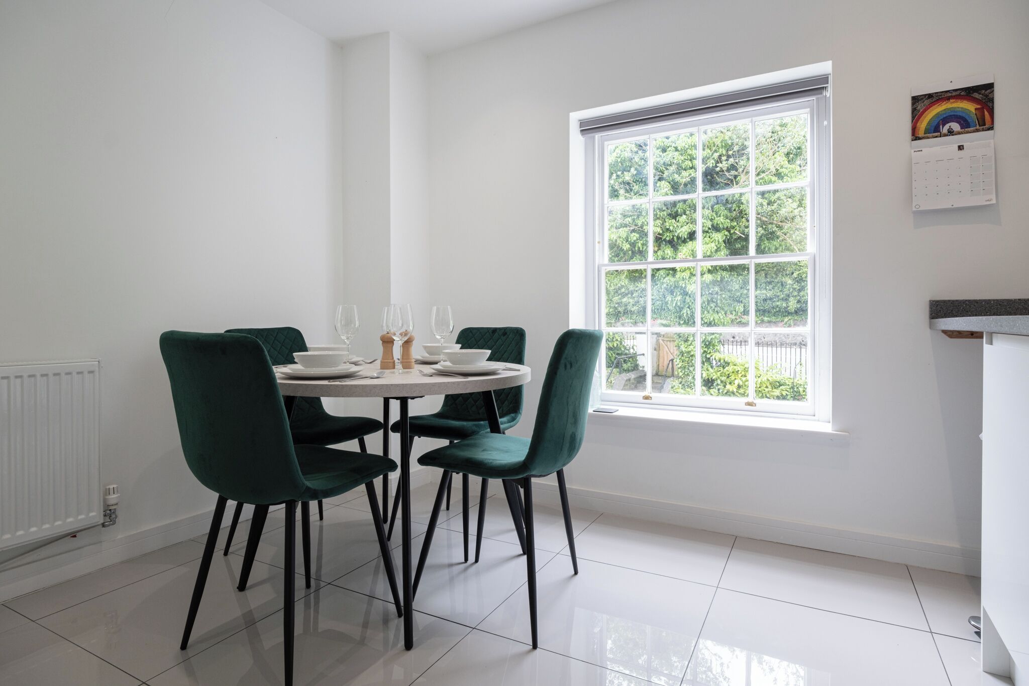 A dining area with a round table, four green chairs, and a window with a view of greenery.