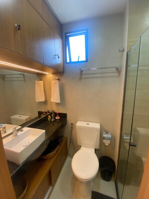 Une petite salle de bain avec un lavabo, des toilettes et une douche, le tout de couleur blanche. Une petite fenêtre laisse entrer la lumière naturelle.