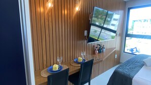 A modern room with a wooden slat wall, a flat-screen TV mounted above a wooden desk, two chairs, a bed with a dark blanket, and a window with a view of the outside.