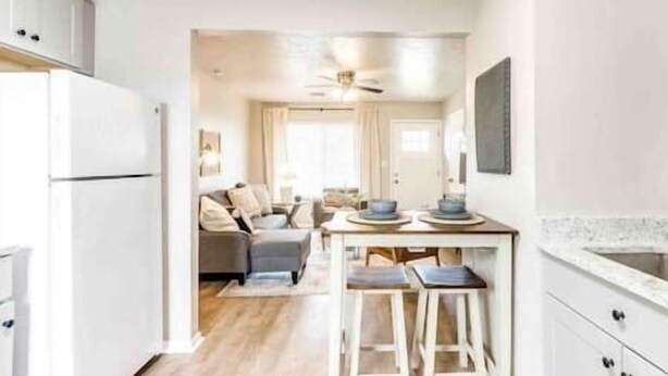 Et kompakt køkken med hvide skabe, et spiseområde med barstole og en stue med sofa og sofabord.