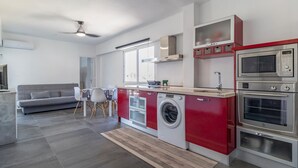A modern kitchen with a built-in washer and dryer, a dining area with a gray sofa, and a large window providing natural light.