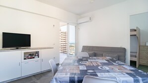 A modern, well-lit room with a flat-screen TV, a dining table with a patterned tablecloth, a grey sofa, and a small kitchenette.